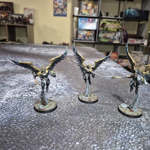 May include: Three miniature figurines of winged warriors. Each figure has detailed armor, a weapon, and large, outstretched wings with gold and blue accents. They are mounted on small, circular bases.