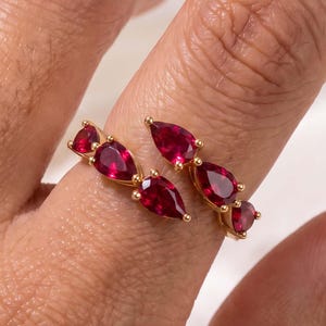 May include: A gold open ring featuring five pear-shaped, deep red gemstones arranged in a bypass style. The band is thin and metallic, with each stone secured in a prong setting, creating a continuous, elegant line across the finger.