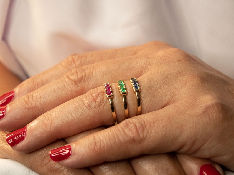 Pu&ograve; includere: Quattro anelli d'oro con pietre preziose di diversi colori. Gli anelli sono impilati su un dito. Le pietre preziose sono rosse, verdi e blu.