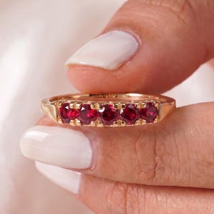 May include: A gold band ring featuring five round-cut, deep red rubies set in a row. The stones are held in place by small metal prongs along the center of the band, creating a continuous line of vibrant red gemstones across the front of the ring.