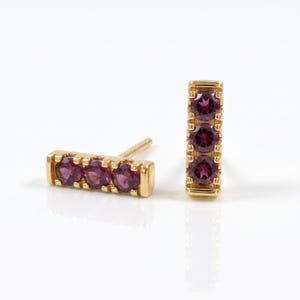 May include: A pair of gold stud earrings, each featuring a rectangular bar design set with three round, deep magenta-colored gemstones. The earrings are positioned with one lying horizontally and the other standing vertically on a white surface.