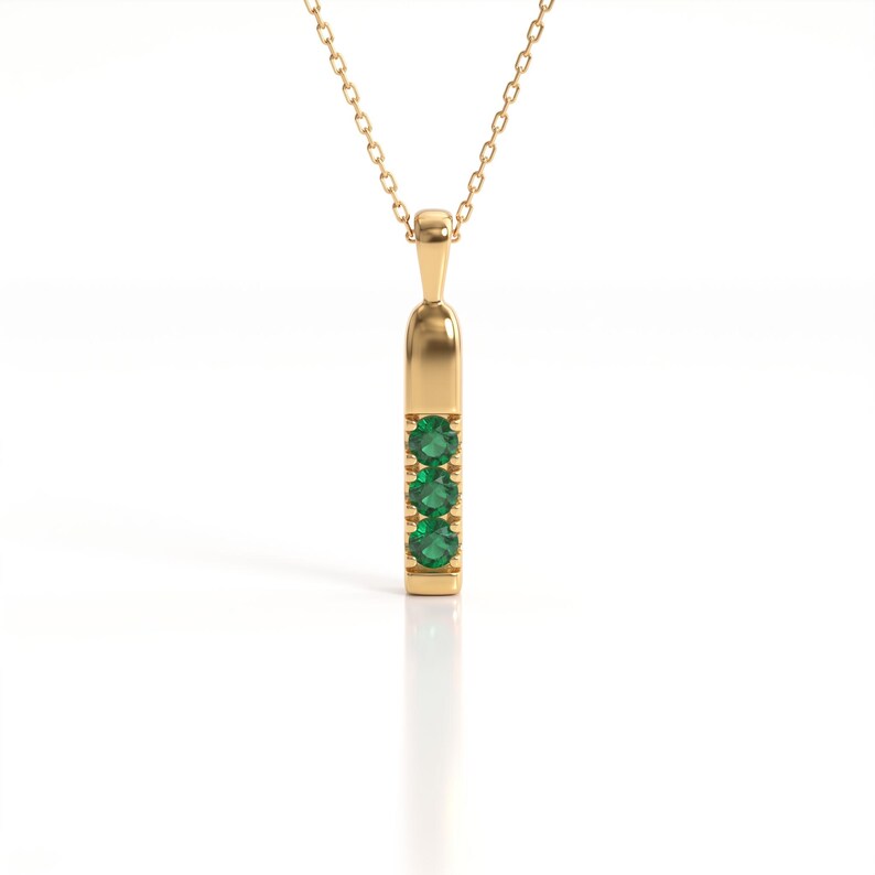 May include: A gold pendant necklace featuring a vertical bar design with three round, vibrant green gemstones set in a row. The pendant is suspended from a delicate gold chain, showcasing a polished metallic finish and a clean, symmetrical structure.