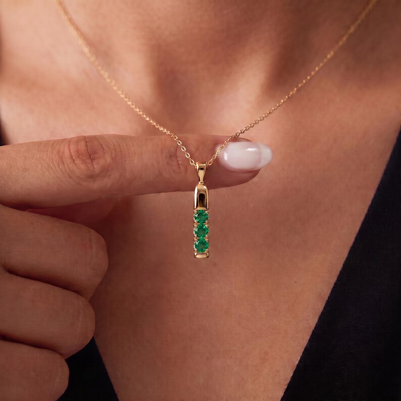 May include: A gold pendant necklace featuring a vertical bar design set with three round, vibrant green gemstones. The pendant hangs from a delicate gold chain, showcasing a polished metal finish and a secure, minimalist setting for the stones.