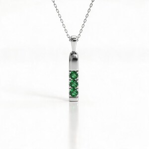May include: A silver-toned pendant necklace featuring a vertical bar design with three round, vibrant green gemstones set in a row. The pendant hangs from a delicate, thin silver-toned chain, creating a clean and balanced look for this jewelry piece.