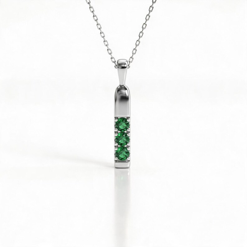 May include: A silver-toned pendant necklace featuring a vertical bar design with three round, vibrant green gemstones set in a row. The pendant hangs from a delicate, thin silver-toned chain, creating a clean and balanced look for this jewelry piece.
