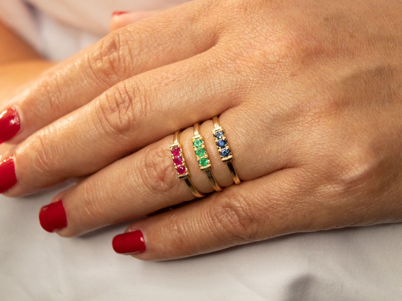 Pu&ograve; includere: Quattro anelli d'oro con tre pietre preziose ciascuno. Gli anelli sono impilati su un dito. Le pietre preziose sono rosse, verdi e blu.