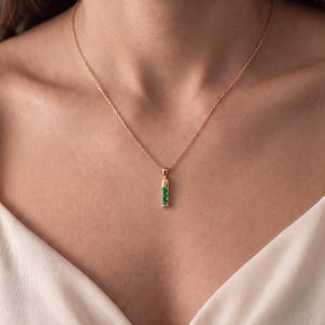 May include: A gold chain necklace featuring a vertical bar pendant. The pendant is crafted from gold and is set with three small, round, vibrant green gemstones arranged in a vertical line, suspended from a delicate, thin gold link chain.