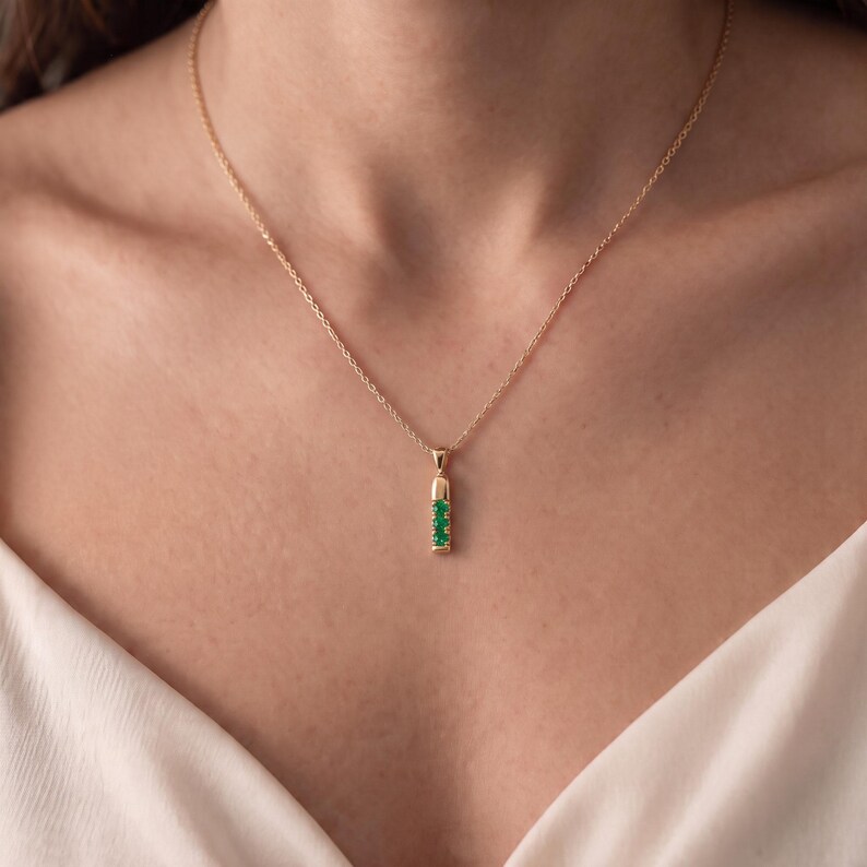May include: A gold chain necklace featuring a vertical bar pendant. The pendant is crafted from gold and is set with three small, round, vibrant green gemstones arranged in a vertical line, suspended from a delicate, thin gold link chain.