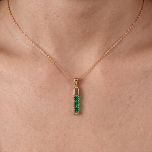 May include: A gold chain necklace featuring a vertical pendant with three round, vibrant green gemstones set in a polished gold metal bar. The pendant hangs from a delicate gold rope chain, creating a simple and elegant piece of jewelry.