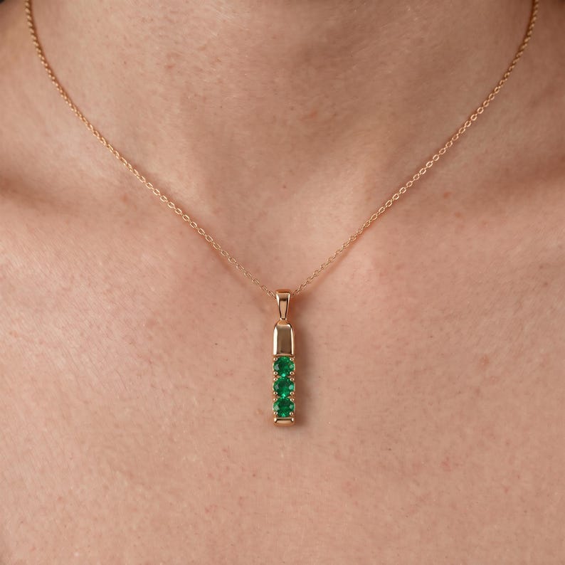 May include: A gold chain necklace featuring a vertical pendant with three round, vibrant green gemstones set in a polished gold metal bar. The pendant hangs from a delicate gold rope chain, creating a simple and elegant piece of jewelry.