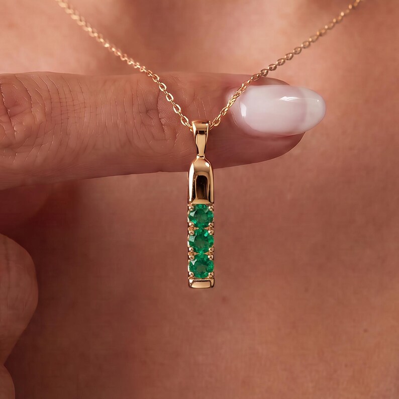 May include: A gold pendant necklace featuring a vertical bar design set with three round, vibrant green emerald gemstones. The pendant is suspended from a delicate gold chain, showcasing a polished metal finish and a secure, minimalist setting.