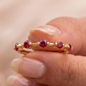 May include: A gold ring featuring five round, vibrant red gemstones set in a horizontal row. The band is thin and polished, with each stone secured by small metal prongs, creating a delicate and symmetrical design across the top of the ring.