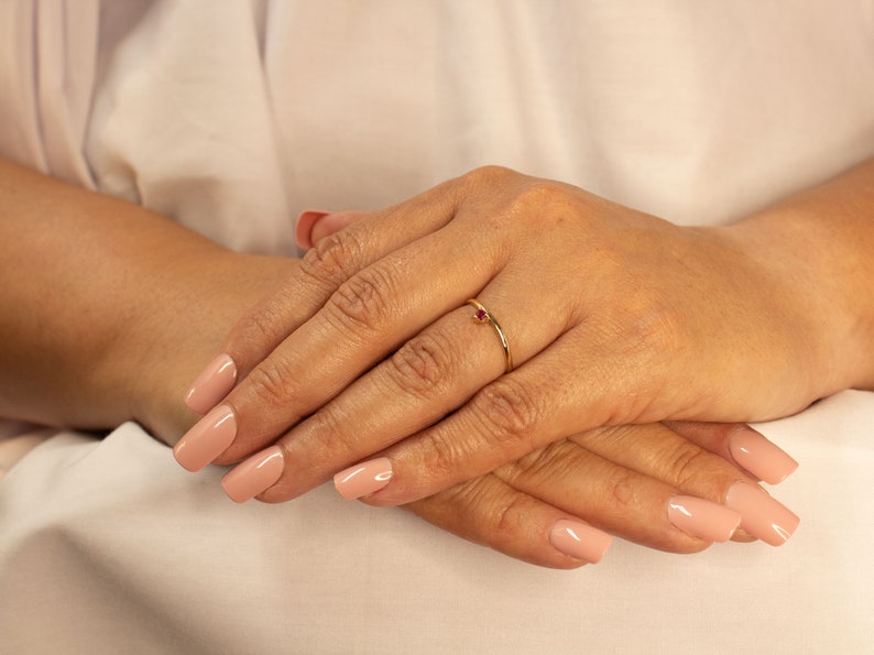 Peut inclure: Une d&eacute;licate bague en or avec une seule pierre pr&eacute;cieuse rouge. La bague est port&eacute;e &agrave; l'annulaire d'une main avec un vernis &agrave; ongles rose clair.