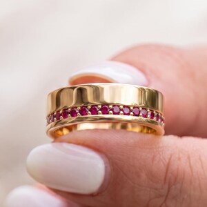 May include: A gold band ring featuring a central horizontal channel set with a continuous row of small, round, vibrant red gemstones. The ring has a polished, smooth gold finish on the upper and lower edges, creating a sleek and refined appearance.