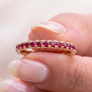 May include: A gold eternity band featuring a continuous row of small, round, deep red rubies set in a shared prong setting. The ring has a polished metal finish and showcases a uniform arrangement of gemstones encircling the entire band.