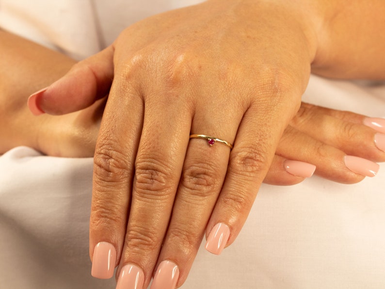 Peut inclure: Une d&eacute;licate bague en or avec une petite pierre pr&eacute;cieuse rouge. La bague est port&eacute;e &agrave; l'annulaire d'une main.