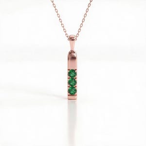 May include: A rose gold pendant necklace featuring a vertical bar design. The pendant is set with three round, vibrant green gemstones arranged in a straight line. The piece hangs from a delicate, matching rose gold chain with small links.
