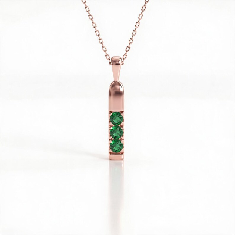 May include: A rose gold pendant necklace featuring a vertical bar design. The pendant is set with three round, vibrant green gemstones arranged in a straight line. The piece hangs from a delicate, matching rose gold chain with small links.