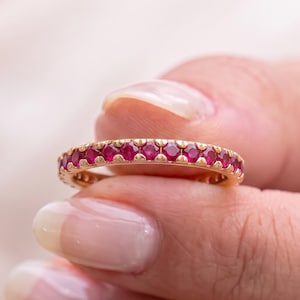 May include: A gold eternity band featuring a continuous row of round, vibrant red rubies set in a polished metal frame. The ring displays a consistent pattern of gemstones secured by small metal prongs, creating a uniform and circular design.