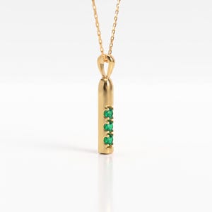 May include: A gold pendant necklace featuring a vertical cylindrical bar design. The pendant is suspended from a delicate gold chain and is set with three small, round, vibrant green gemstones arranged in a vertical line along the front surface.