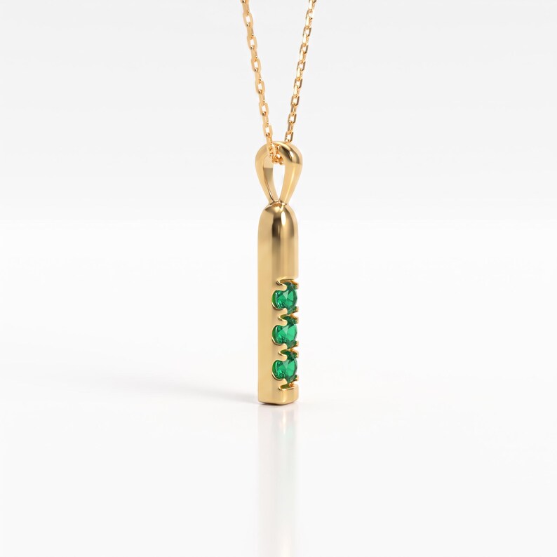 May include: A gold pendant necklace featuring a vertical cylindrical bar design. The pendant is suspended from a delicate gold chain and is set with three small, round, vibrant green gemstones arranged in a vertical line along the front surface.