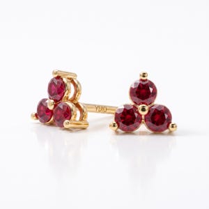 May include: A pair of gold stud earrings, each featuring three round-cut red rubies arranged in a triangular cluster. The metal post of one earring is clearly visible and features the engraved text YUKI, indicating the brand or maker mark.