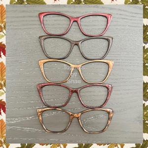 May include: Five pairs of cat-eye glasses in different colors and patterns. The glasses are arranged in a stack, with the top pair being red, followed by brown, gold, red, and gold.