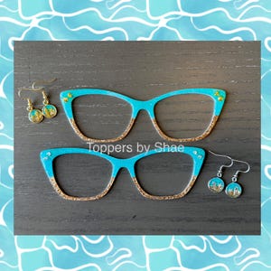 May include: A pair of turquoise blue cat eye glasses with gold glitter accents and matching earrings with gold glitter accents. The glasses and earrings are on a wooden surface. "Toppers by Shae" is written on the surface.