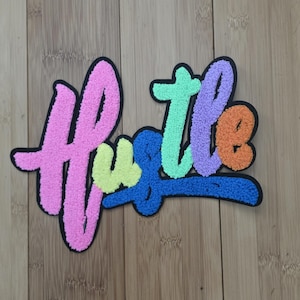 May include: A multi-colored chenille patch featuring the word Hustle in a script font. The letters are rendered in bright pink, lime green, blue, mint green, purple, and orange, all outlined in a thick black border with a textured surface.