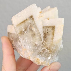 May include: A cluster of off-white, crystalline mineral formations with a layered, geometric structure. The mineral has a translucent quality with subtle brown and tan inclusions. The specimen is held in a hand against a gray background.