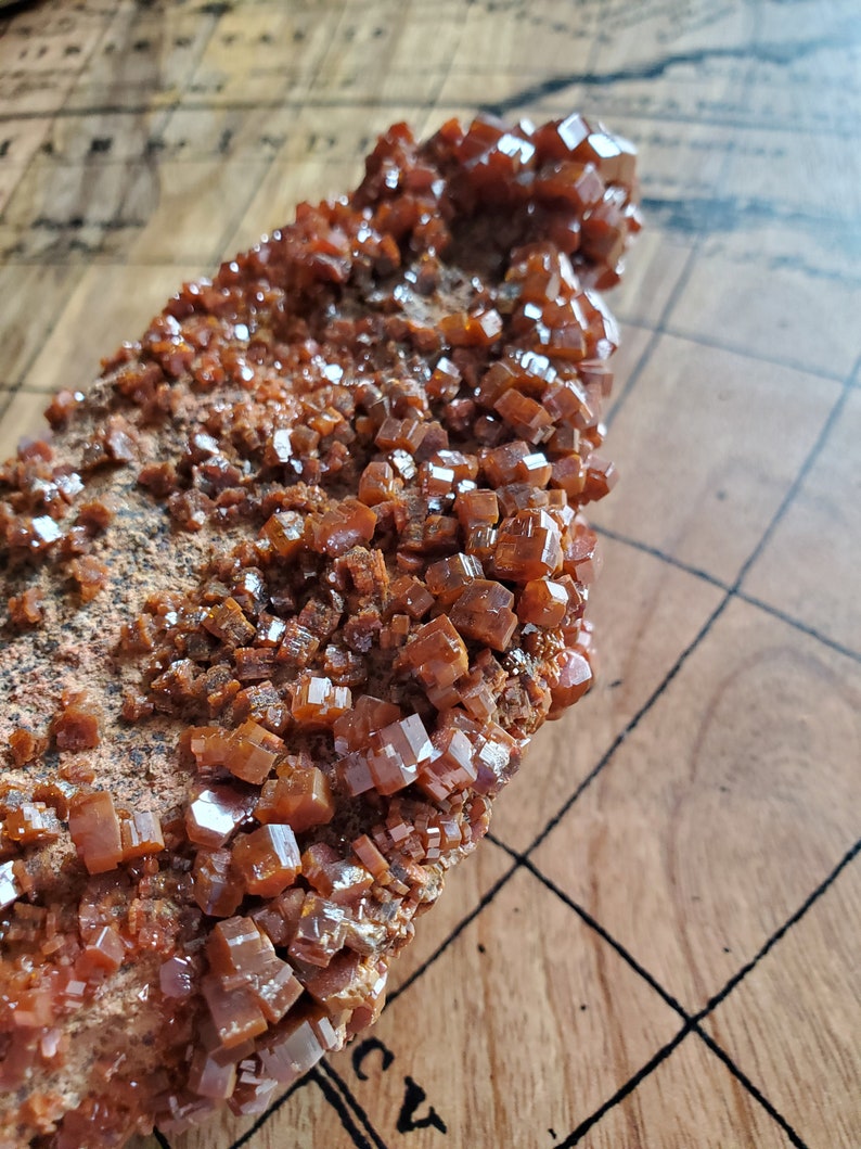 May include: A close-up of a rough, brown rock covered in small, shiny, orange crystals. The crystals are clustered together and have a cubic shape.