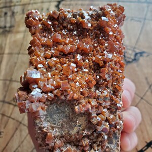 May include: A cluster of orange-brown crystals on a dark brown rock. The crystals are small and have a shiny, almost metallic, appearance.