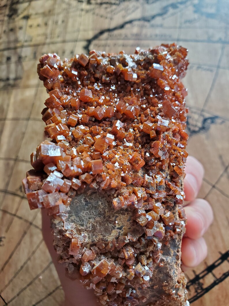 May include: A cluster of orange-brown crystals on a dark brown rock. The crystals are small and have a shiny, almost metallic, appearance.