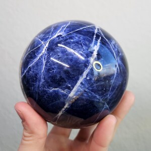 May include: A polished, spherical sodalite gemstone. The sphere is a deep blue color with white veining throughout, creating a marbled effect. The gemstone is held in a hand, showcasing its smooth, reflective surface. A decorative item.