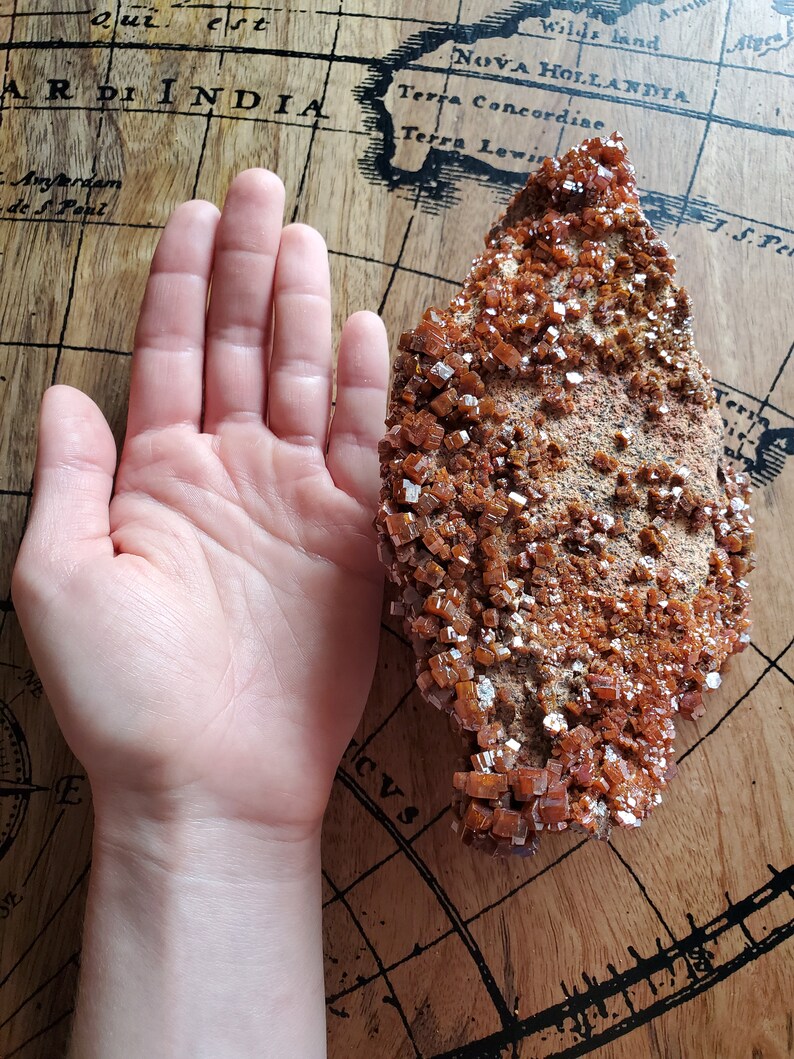 May include: A large, rough, orange-brown rock formation with a cluster of small, shiny, orange crystals. The rock is sitting on a wooden surface.