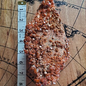 May include: A large, rough, orange-brown rock covered in small, shiny, orange crystals. The rock is about 7 inches long and is sitting on a wooden surface with a map printed on it.