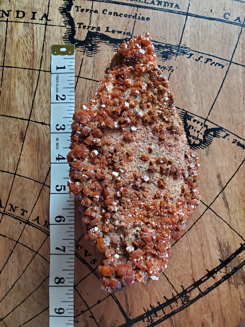 May include: A large, rough, orange-brown rock covered in small, shiny, orange crystals. The rock is about 7 inches long and is sitting on a wooden surface with a map printed on it.