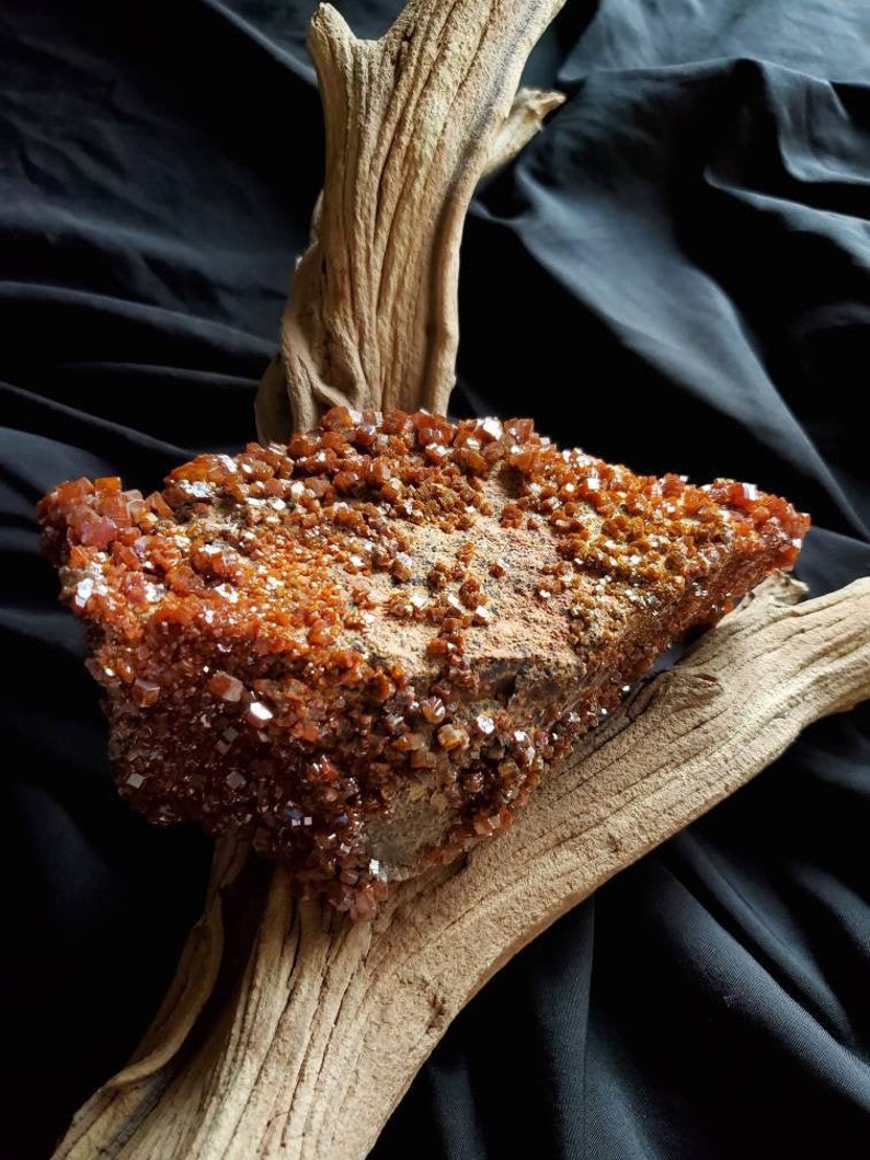 May include: A large, rough, orange-brown rock covered in small, sparkling crystals. The rock is resting on a piece of driftwood.