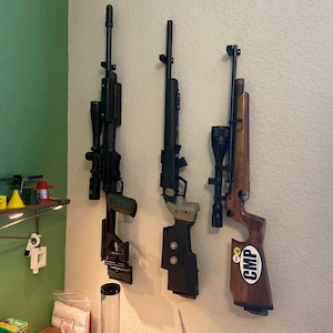 Könnte beinhalten: Drei Gewehre an einer weißen Wand montiert. Die Gewehre sind schwarz und braun mit angebrachten Zielfernrohren. Das Gewehr rechts hat einen Aufkleber mit der Aufschrift "CMP".