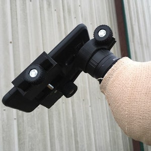 May include: A black smartphone attached to a telescope with a black phone holder. The phone holder has two knobs for adjusting the phone's position. The telescope is covered in a beige fabric.