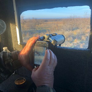 May include: A person is taking a picture of a deer through a window with a smartphone. The deer is visible on the phone screen. The person is holding binoculars in their other hand.