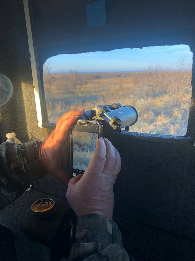 May include: A person is taking a picture of a deer through a window with a smartphone. The deer is visible on the phone screen. The person is holding binoculars in their other hand.