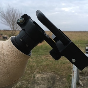 May include: A black telescope with a white fabric cover attached to the base. A black smartphone is mounted to the telescope using a black bracket. The telescope is in a field with dry grass and a cloudy sky.