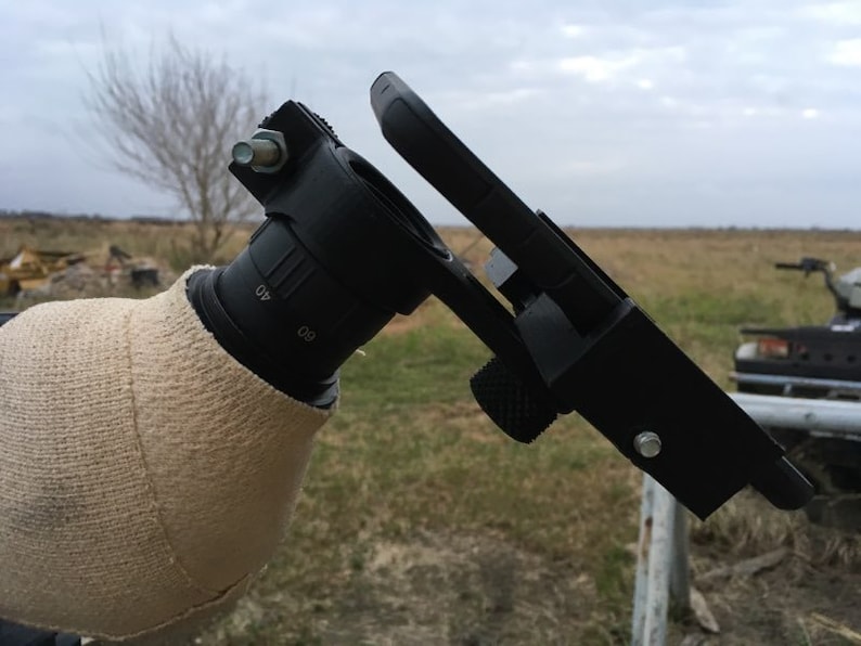 May include: A black telescope with a white fabric cover attached to the base. A black smartphone is mounted to the telescope using a black bracket. The telescope is in a field with dry grass and a cloudy sky.