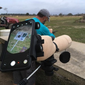 May include: A black smartphone is mounted to a beige telescope scope. The phone is displaying a target with a bullseye. The scope is mounted on a tripod. The phone has a red recording button.
