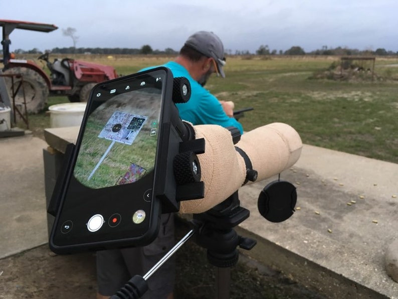 May include: A black smartphone is mounted to a beige telescope scope. The phone is displaying a target with a bullseye. The scope is mounted on a tripod. The phone has a red recording button.
