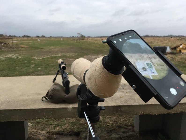 May include: A black rifle scope mounted on a tripod with a smartphone attached to the scope using a phone holder. The phone is displaying a target with a bullseye.