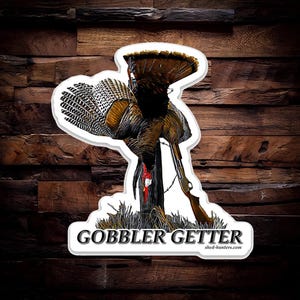 Può includere: Un adesivo con un'illustrazione dettagliata di un tacchino con un fucile. Il tacchino è marrone, nero e bianco, con le penne della coda spiegate. Il testo "GOBBLER GETTER" è in basso.