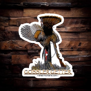 May include: A sticker featuring a detailed illustration of a wild turkey with its wings spread, a shotgun, and the text "GOBBLER GETTER - shed-hunters.com - ".