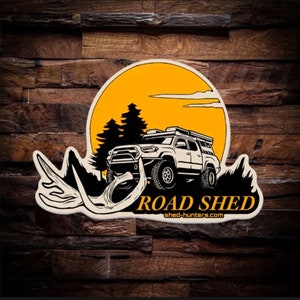 May include: A white pickup truck with a roof top tent drives through a forest with a yellow sun and antlers in the foreground. The text "ROAD SHED" and "shed-hunters.com" are below the truck.